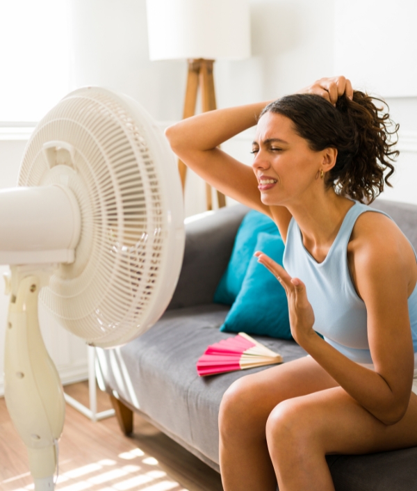 Das Bild zeigt eine Dame die auf dem Sofa vor einem Ventilator sitzt und sich abkühlt.