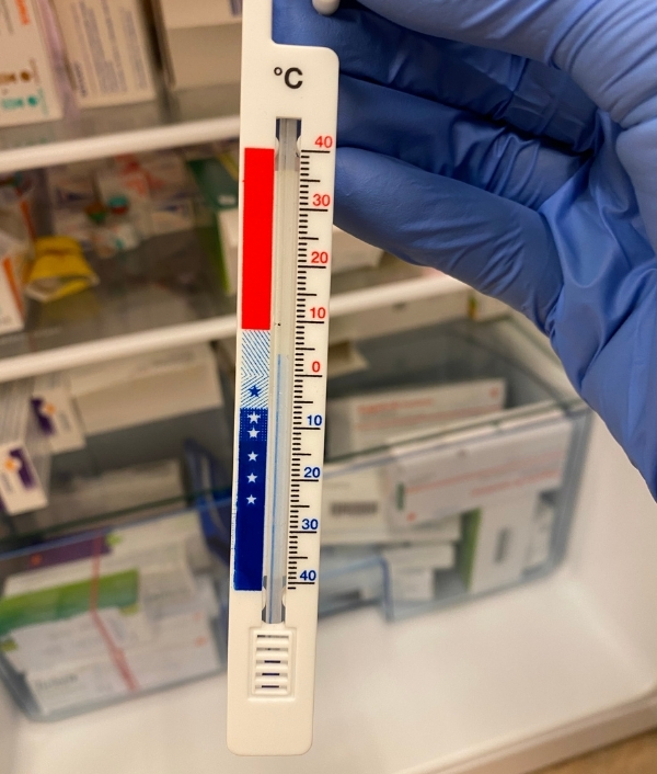 Das Bild zeigt ein Thermometer das von einer Person mit blauem Latexhandschuh gehalten wird. Im Hintergrund sind Medikamente in einem Kühlschrank zu sehen.