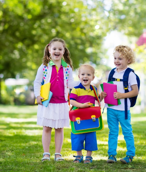Das Bild zeigt drei Kinder zum Schulstart. Alle drei Kinder lachen und befinden sich in einem Park.