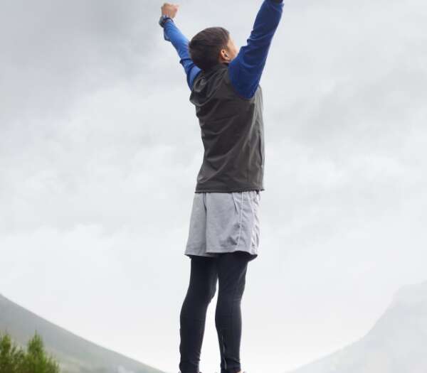 © Canva Das Bild zeigt eine Person im sportlichen Gewand auf einem Berg. Die streckt die Hände in die höhe und strotzt vor Vitalität.