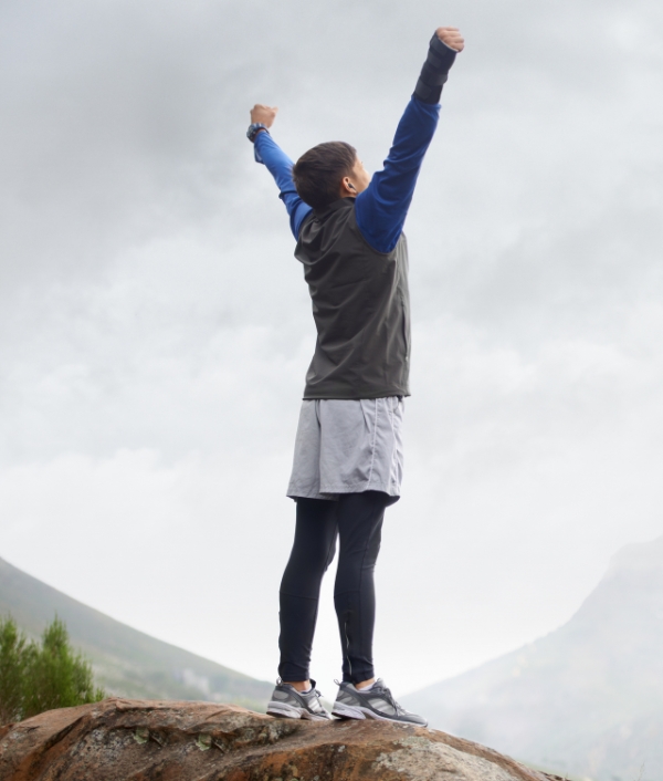 Das Bild zeigt eine Person im sportlichen Gewand auf einem Berg. Die streckt die Hände in die höhe und strotzt vor Vitalität.
