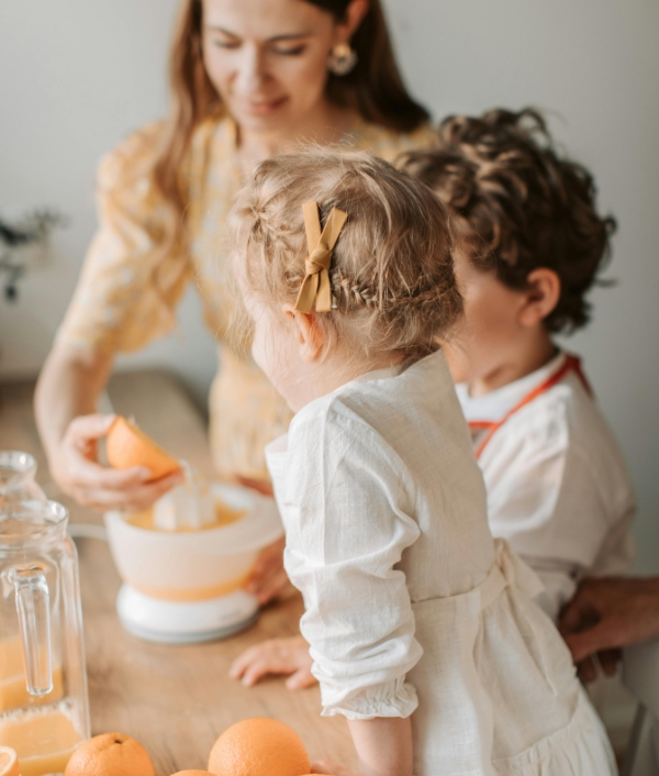 Das Bild zeigt wie eine Mutter ihren beiden Kindern Orangen presst für eine bessere Vitaminzufuhr.